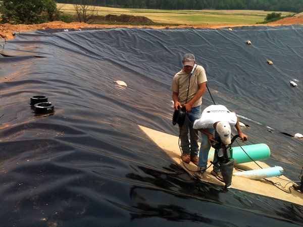 Two men using tools to weld together a black plastic liner in a large outdoor construction site.
