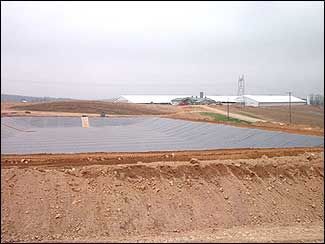 A large, open pond covered with dark material in front of a white industrial building on a cloudy day.