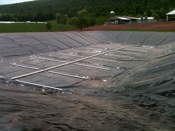 A lined pond with a grid of pipes, set in a rural landscape with a barn and hills in the background.
