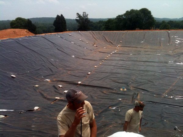 Two men inspecting a large, black liner in an earthen basin, possibly a pond or reservoir. The liner is secured with white weights.