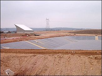 A large lined pit in a construction site, possibly for water or waste storage. A building and power lines are visible in the background.