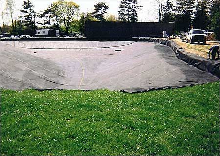 Black liner in a pond basin, ready for water, bordered by green grass and construction materials.