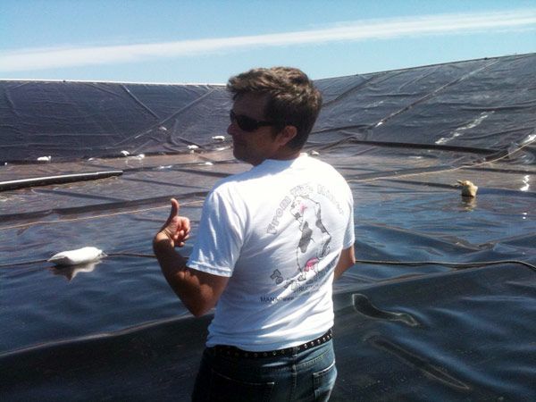 Man in white shirt and sunglasses giving a thumbs up, standing near a large, black water containment structure, blue sky in background.