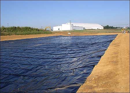 A large black liner covers a shallow, rectangular pit in an outdoor construction site. A white building is in the background under a blue sky.