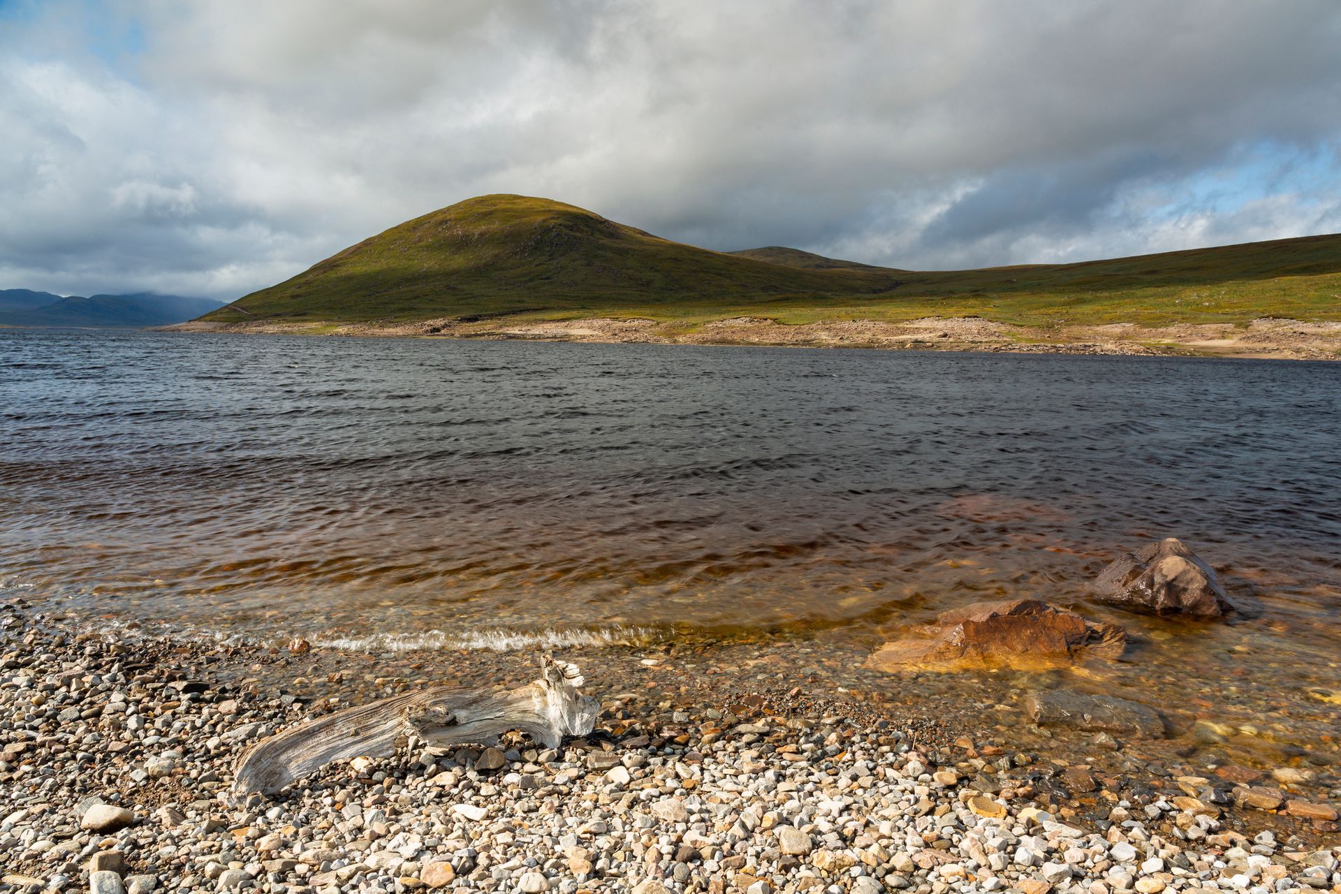 Rocky shoreline of a lake with a green hill under a cloudy sky. Brown and blue tones dominate the landscape.