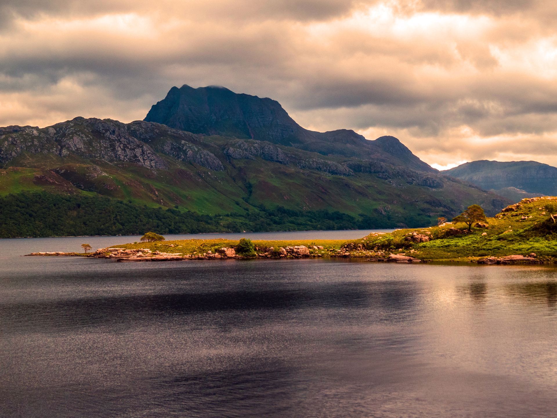 Mountain rising above a lake on an overcast day; brown and green grassy terrain with dark water and sky.