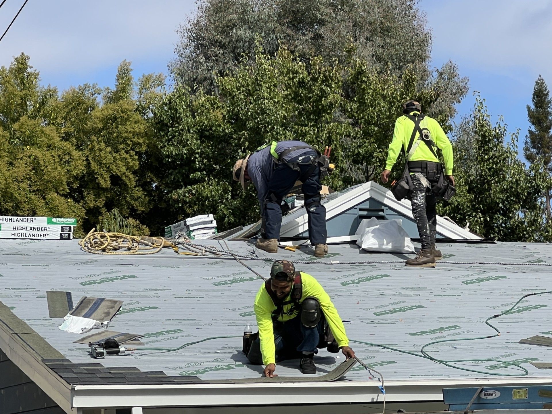 Roofers working on a residential roof, wearing safety gear. Blue sky, green trees, and blue trim are visible.
