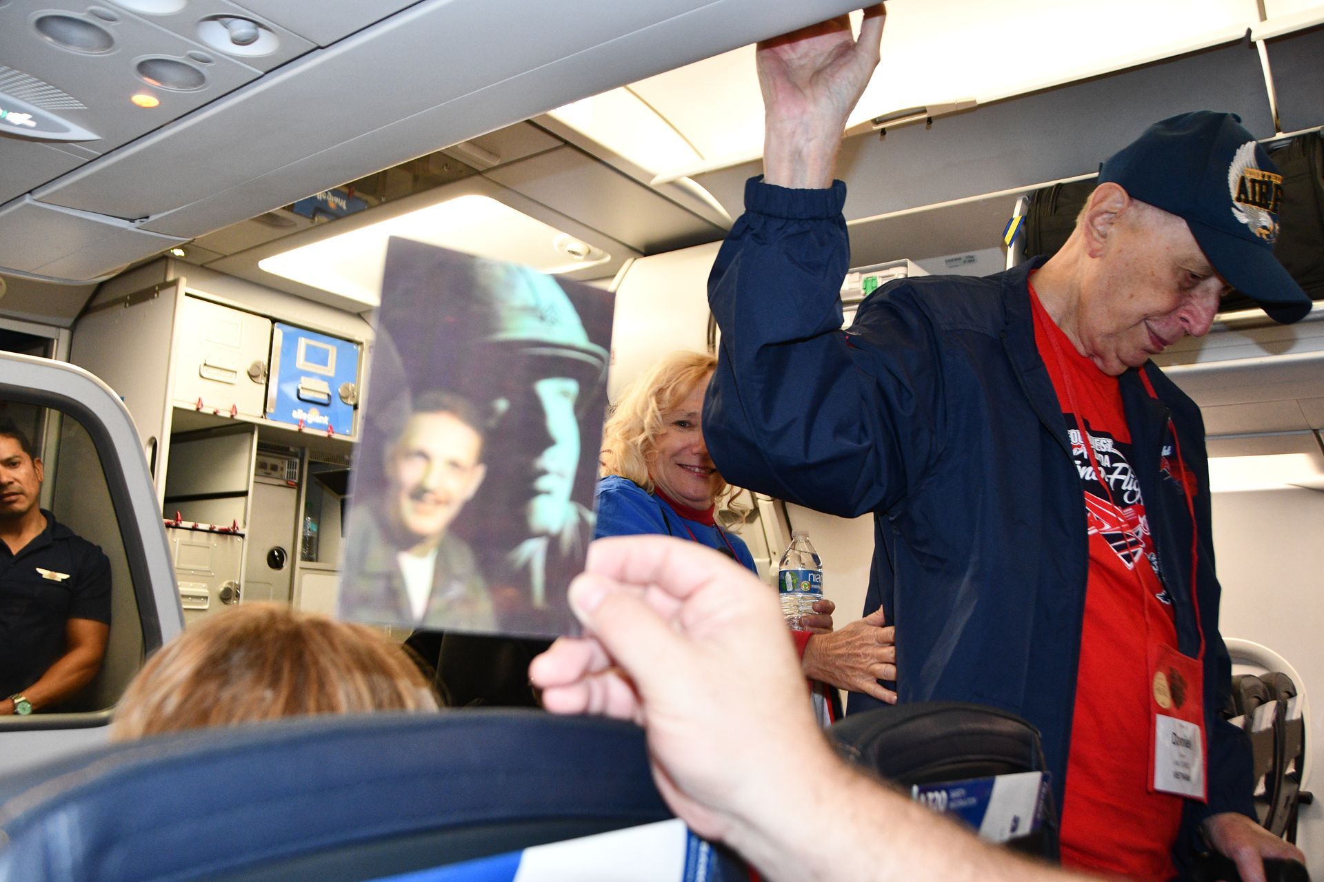 Man in blue hat on airplane, holding a photo; others nearby smiling.