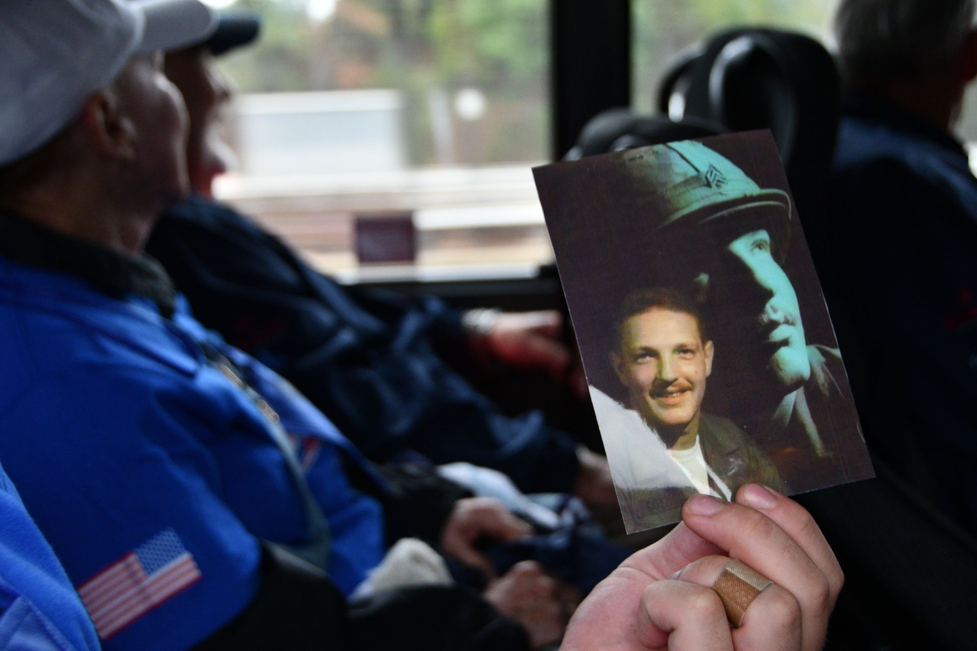 Person holds photo of a young soldier, likely a veteran on a bus. Other veterans are in the background.
