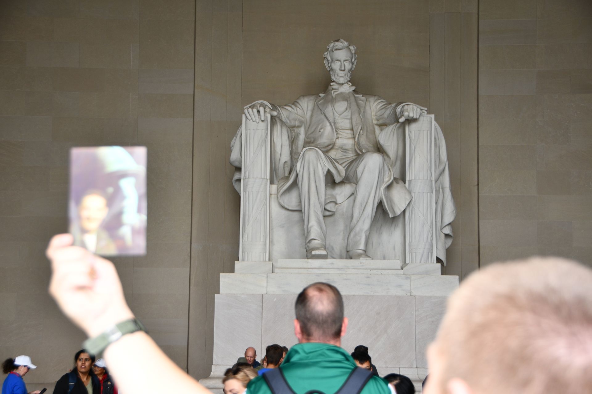 Statue of Lincoln at the Lincoln Memorial. Person holds up a photo of Lincoln. Tourists in background.