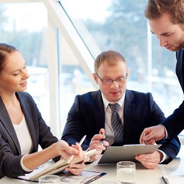 Three business people review documents at a table; one points at a clipboard, window in background.