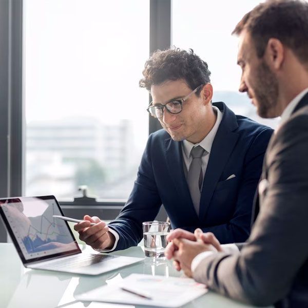Two men in suits reviewing data on a laptop in a bright office. One points at the screen.