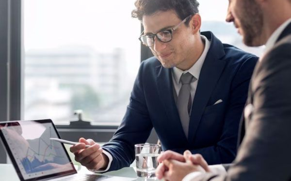 Two men in suits reviewing data on a laptop in an office. One points at the screen.