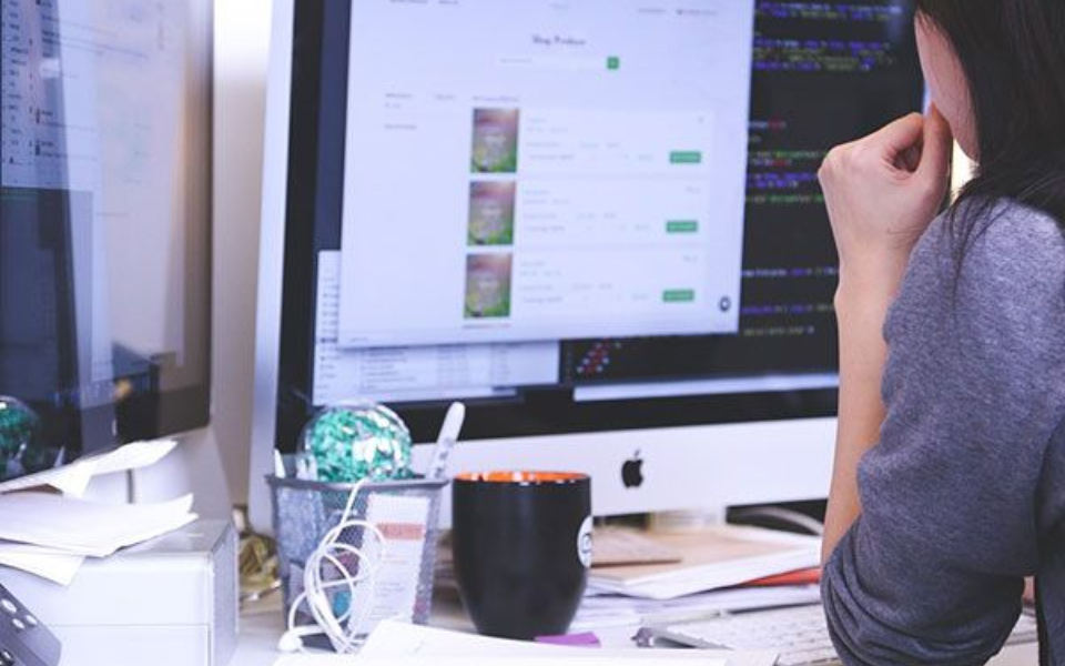 Woman at computer, reviewing web page with product images, hand to face, at desk with coffee cup and office supplies.
