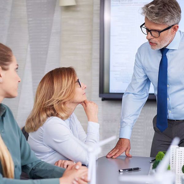A man in a blue shirt gestures at a screen, talking to two women at a conference table.