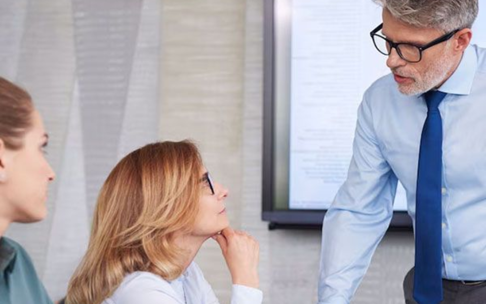 Man in glasses speaking to two women in an office; all looking at a screen.