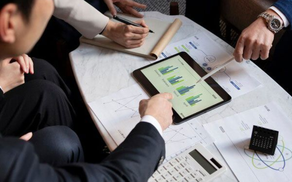 Businesspeople review charts on a tablet, with a calculator and notepad on a table.