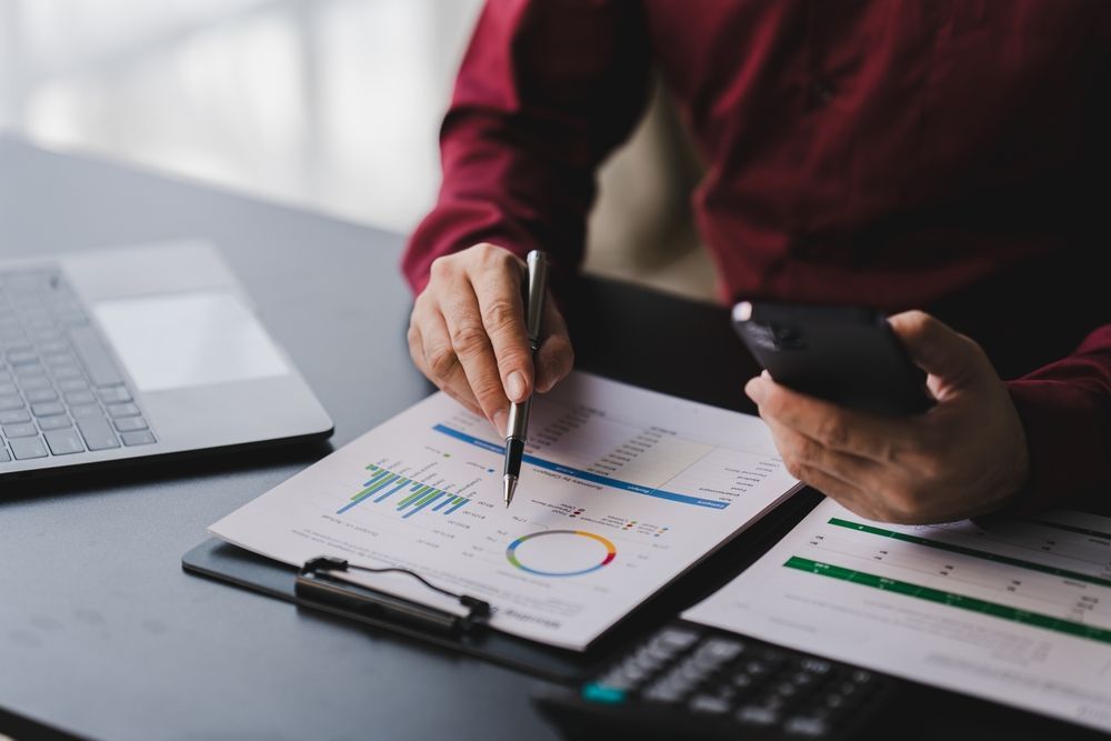 Person analyzing graphs on paper with a pen, holding a phone, laptop and calculator nearby.