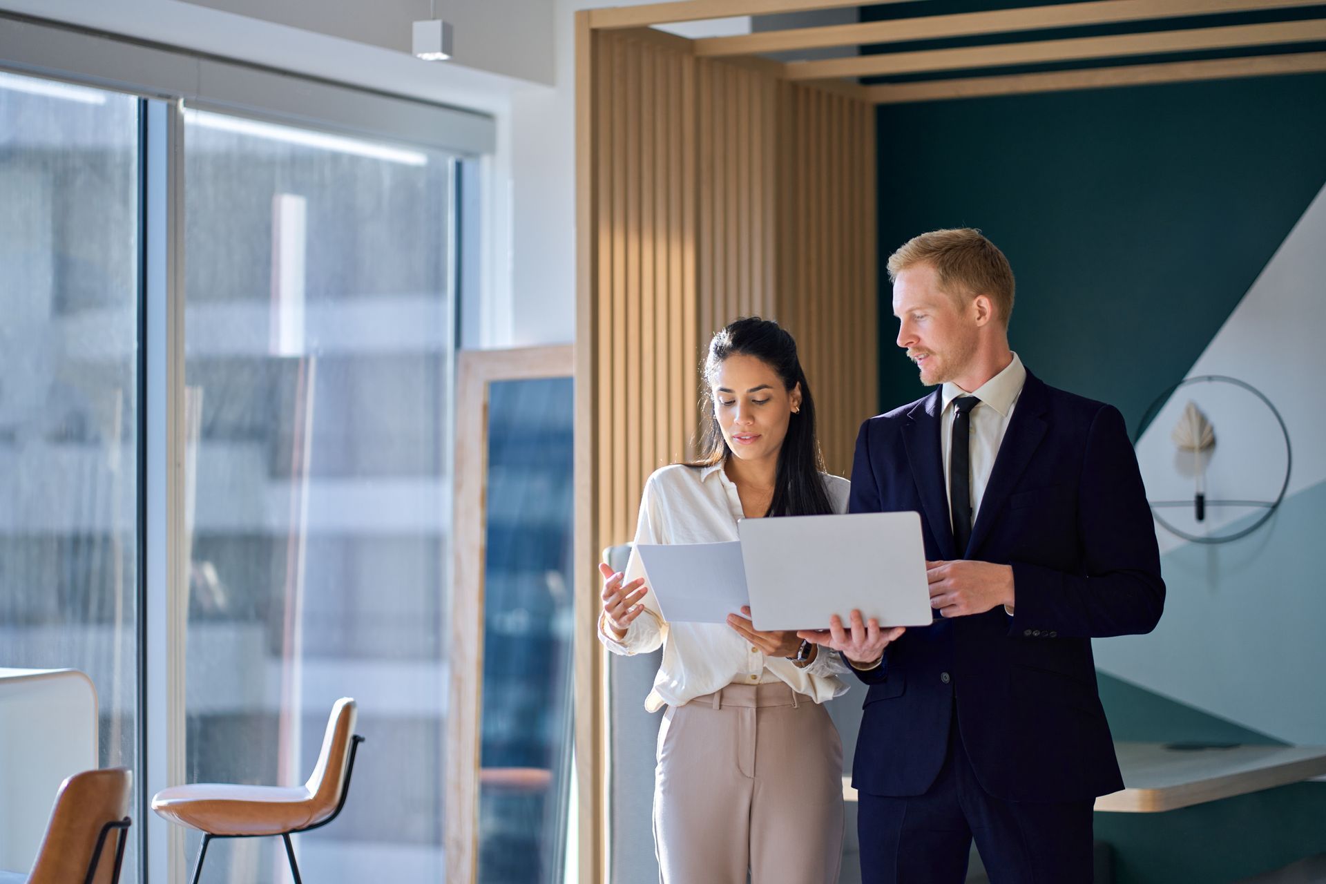 Two businesspeople, a woman and a man, review documents together in an office with a window.