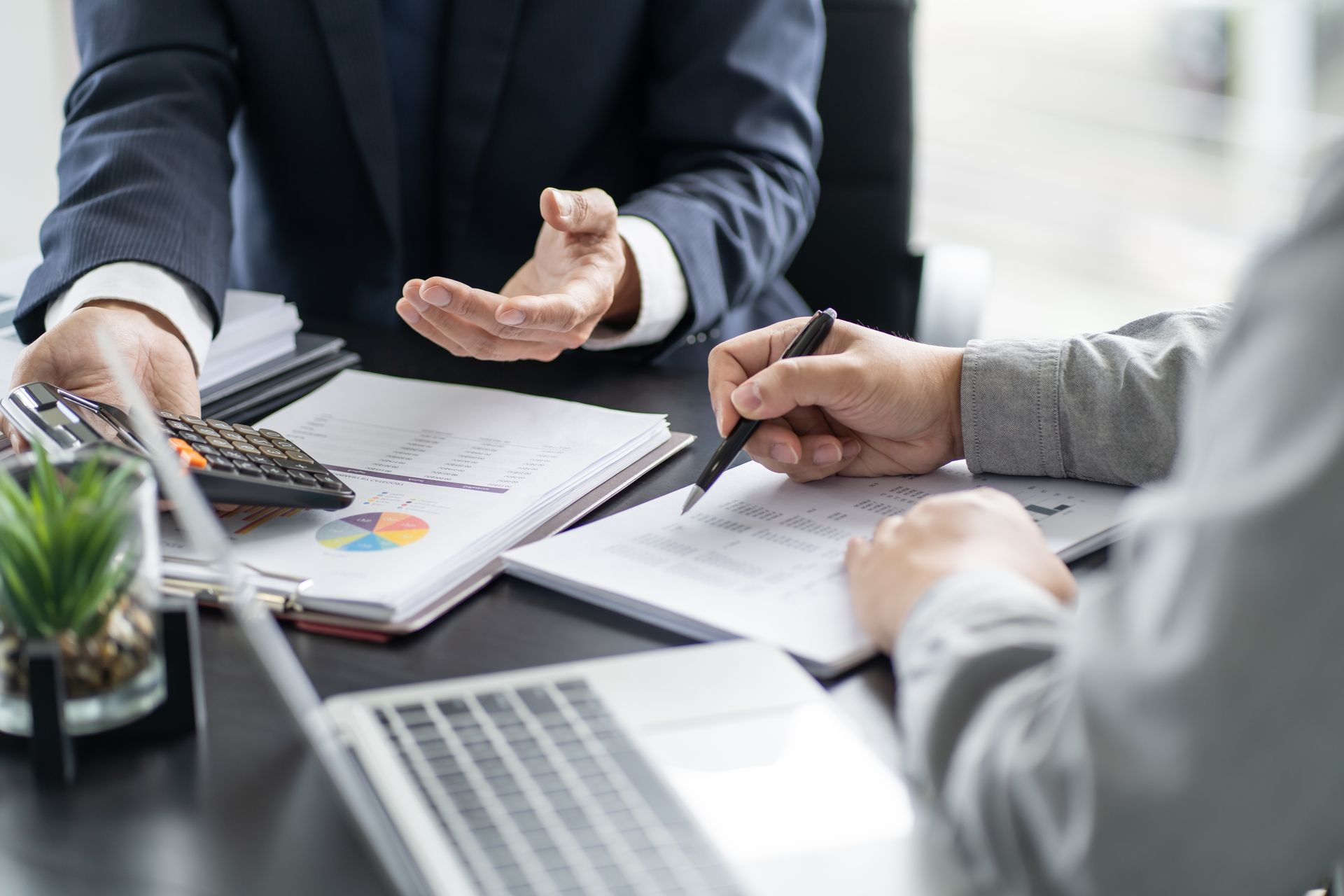 Two people at a desk review financial documents. One gestures, the other writes. Laptop and calculator in view.