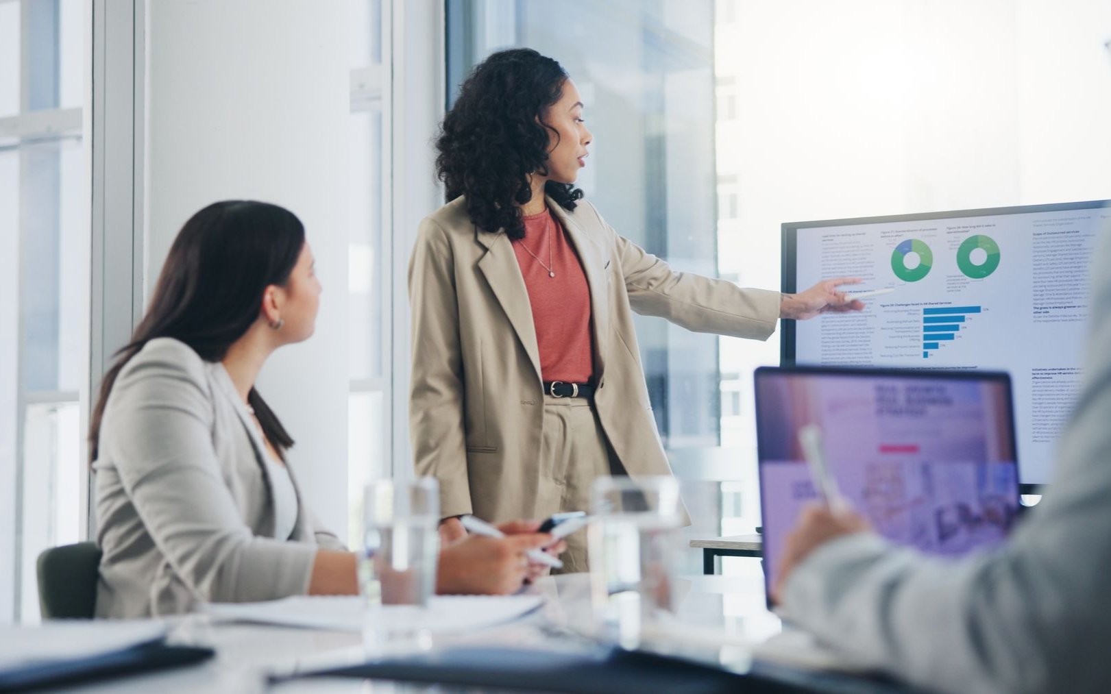Woman in blazer presenting data on screen to colleagues in a conference room.