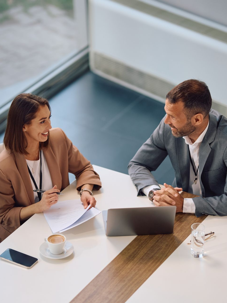 Two people in business attire at a table, smiling and looking at each other, with laptop and documents.