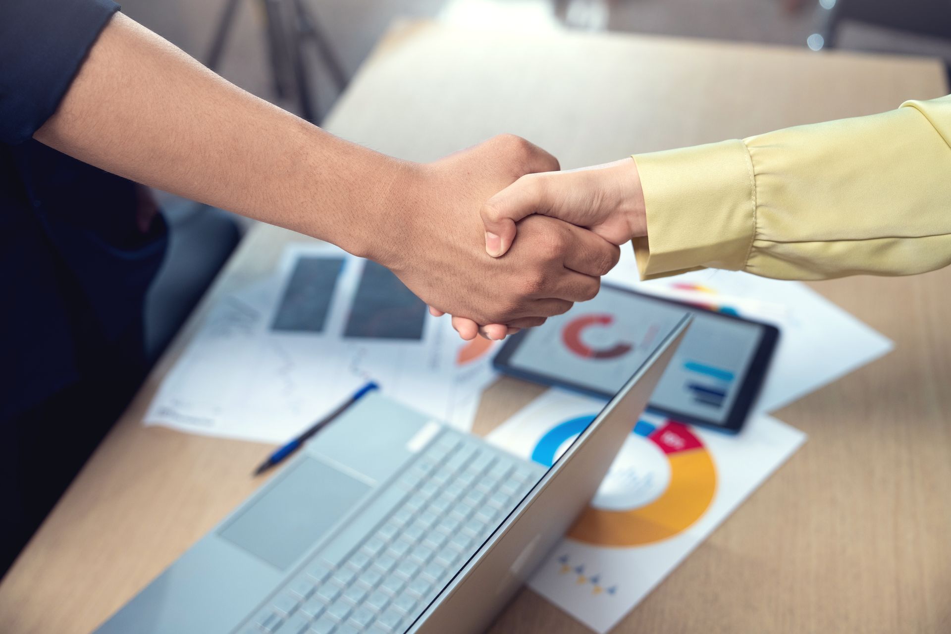 Two people shaking hands over a desk with a laptop, tablet, and charts.