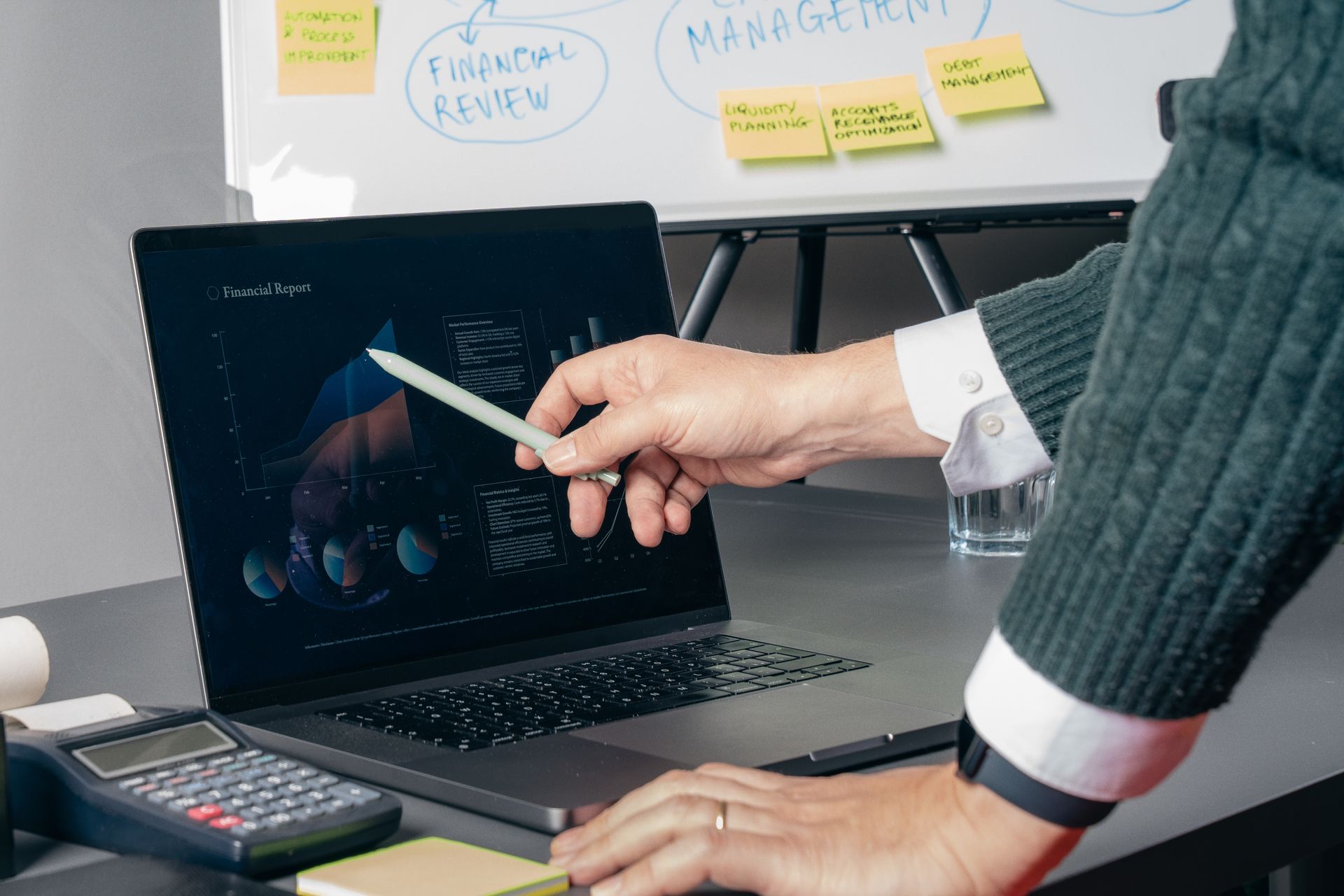 Person pointing to data on a laptop screen; whiteboard with notes in background.
