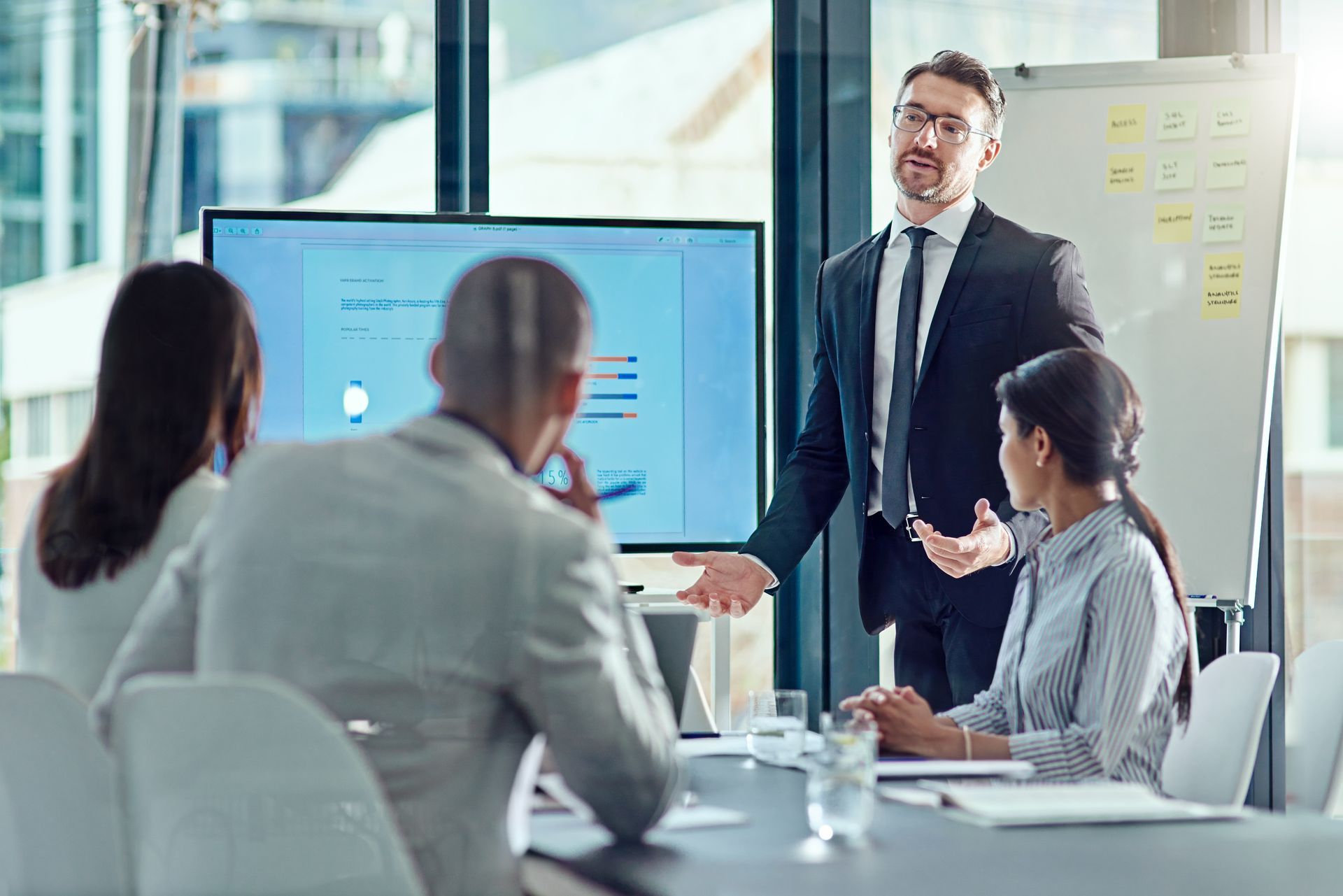 Man in suit presenting in a meeting, pointing at a screen displaying data. Other business people are seated at a table.
