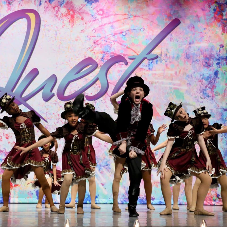 A group of people are dancing on a stage in front of a sign that says quest