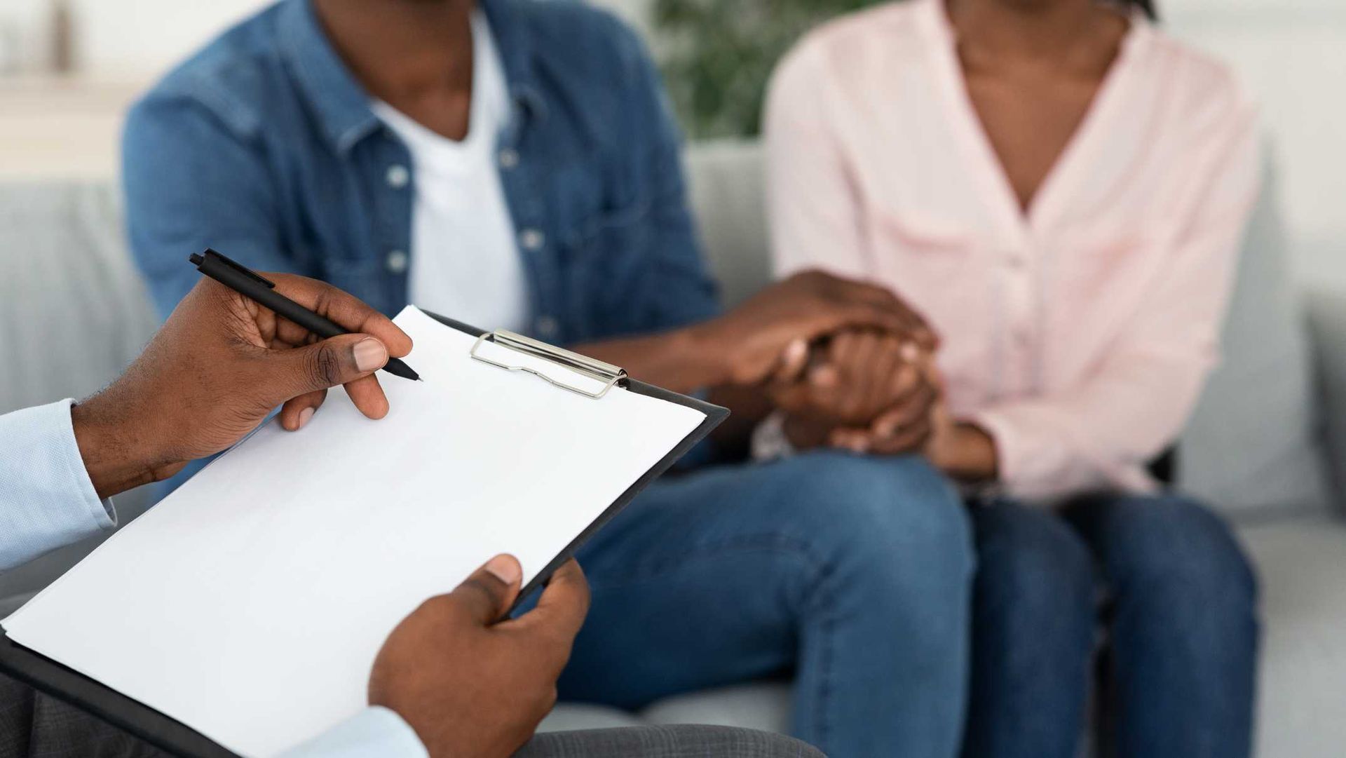 Therapist writing on a clipboard while couple holds hands on a couch.