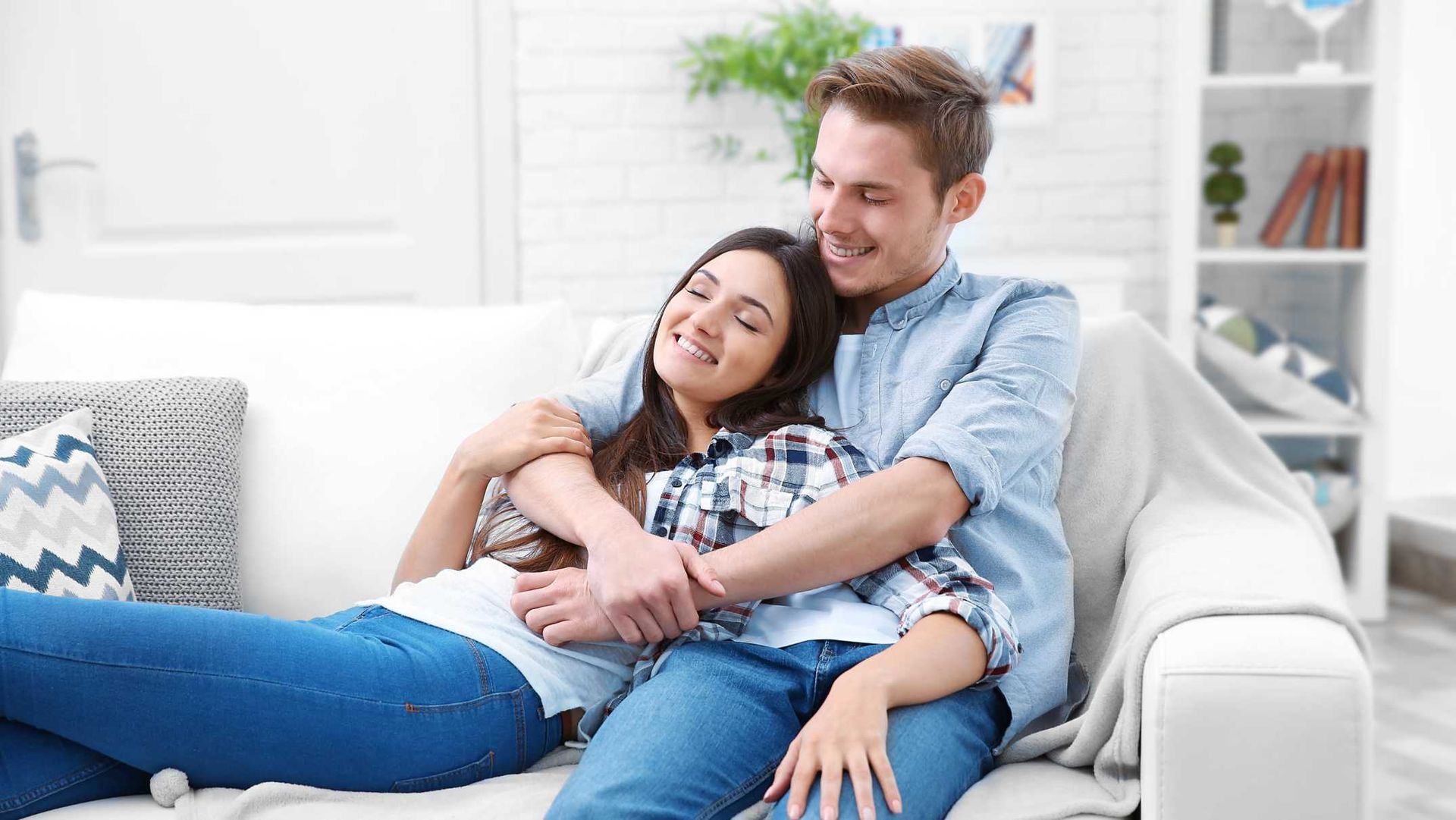 Couple embracing on a white couch; man smiling, woman with eyes closed; bright living room setting.