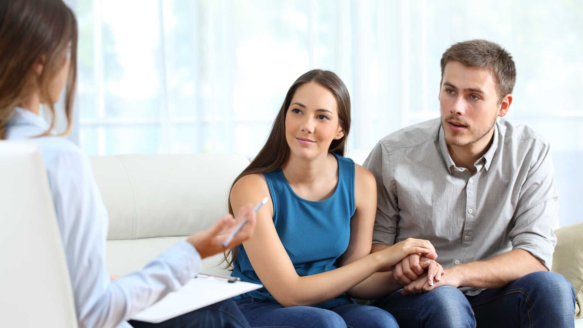 A couple holds hands while listening to a therapist. They are seated on a couch in a bright room.