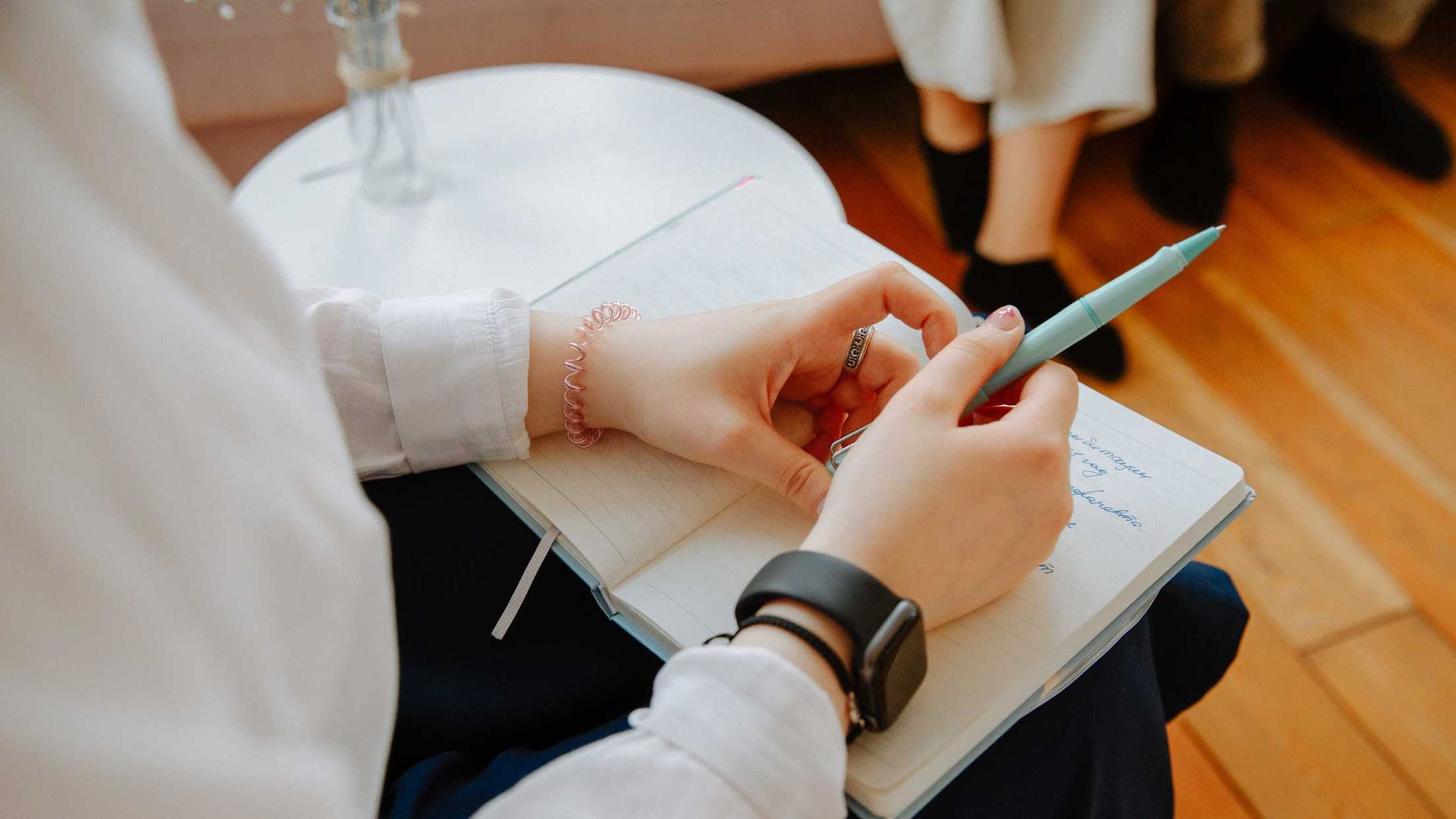 Person writing in a notebook, holding a pen. A smartwatch and bracelet are visible.