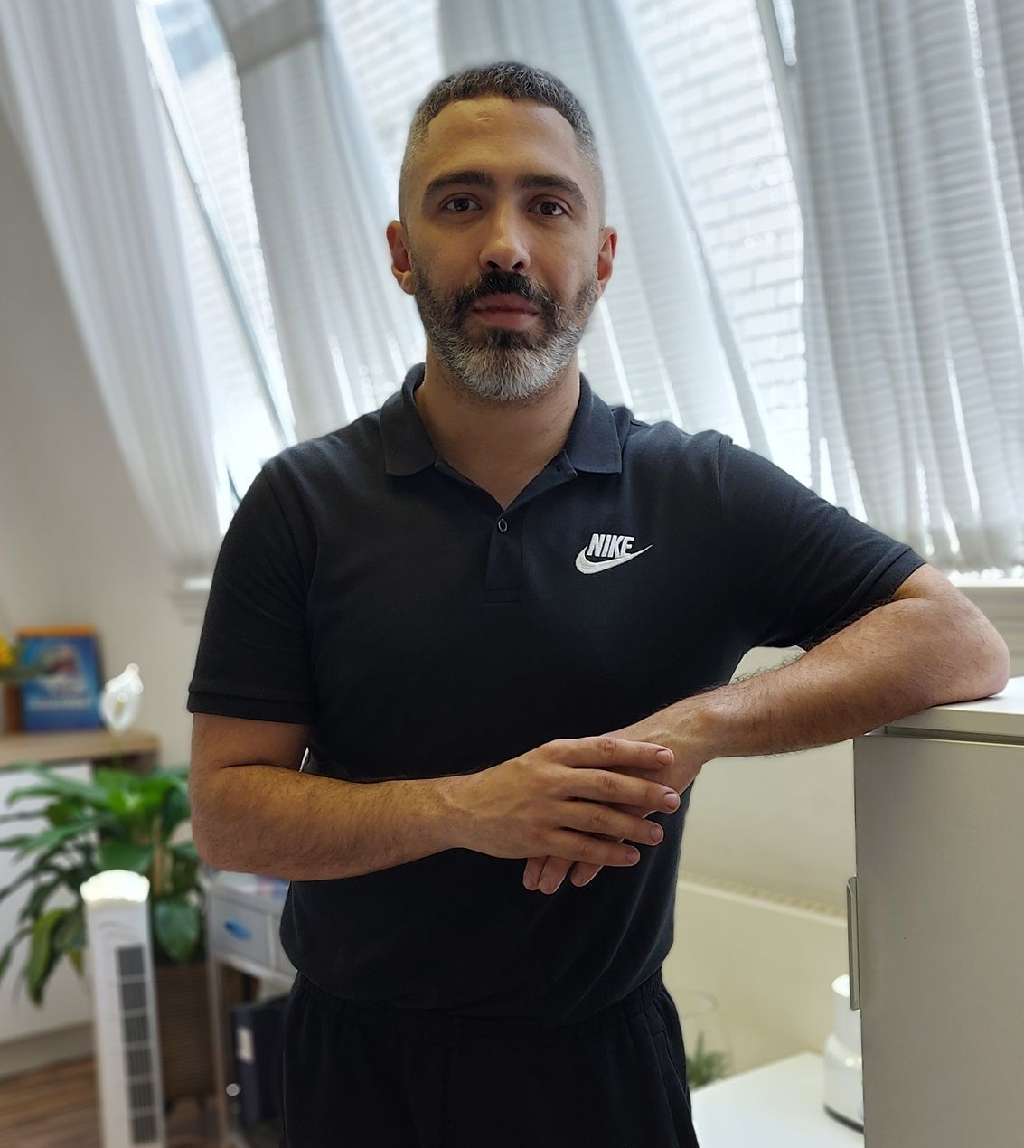 Manil, a Sports Therapist at R P Sports Injuries and Wellness Clinic, is standing confidently in the clinic wearing a black Nike polo shirt, with natural light coming through large windows in the background.