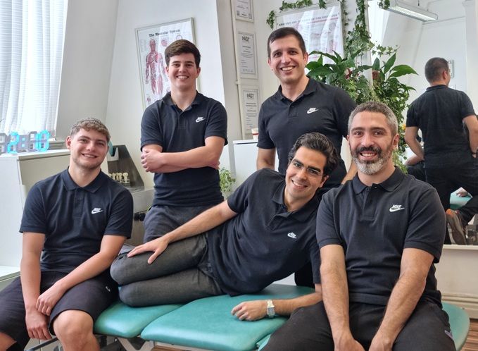 A group of five expert sports therapists at R P Sports Injuries & Wellness Clinic smiling together in a treatment room, seated and standing around a physiotherapy bed, wearing matching black uniforms, with clinic equipment and anatomical models in the background.