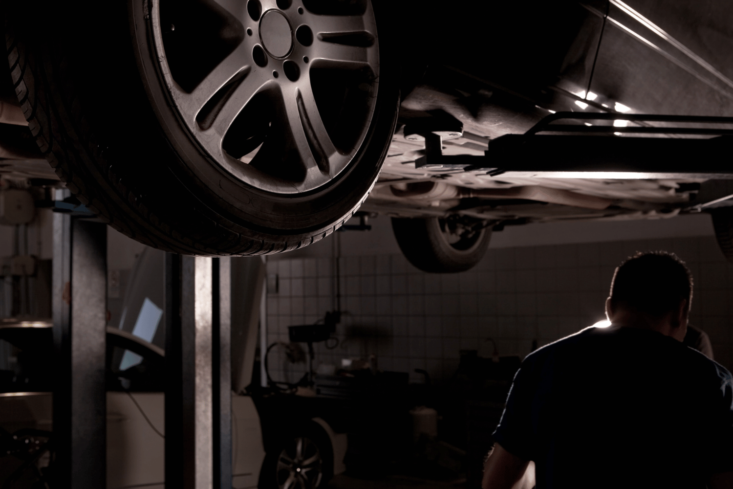 A car lifted in a repair shop; mechanic at work, dark environment.