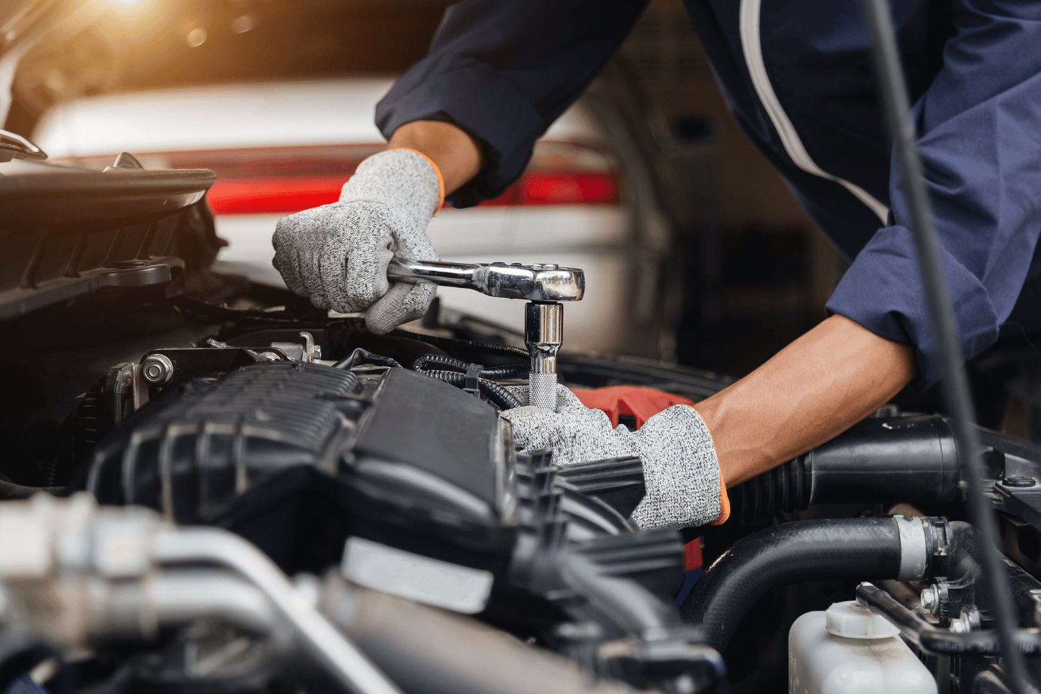 Mechanic wearing gloves, using a wrench on a car engine under the open hood.