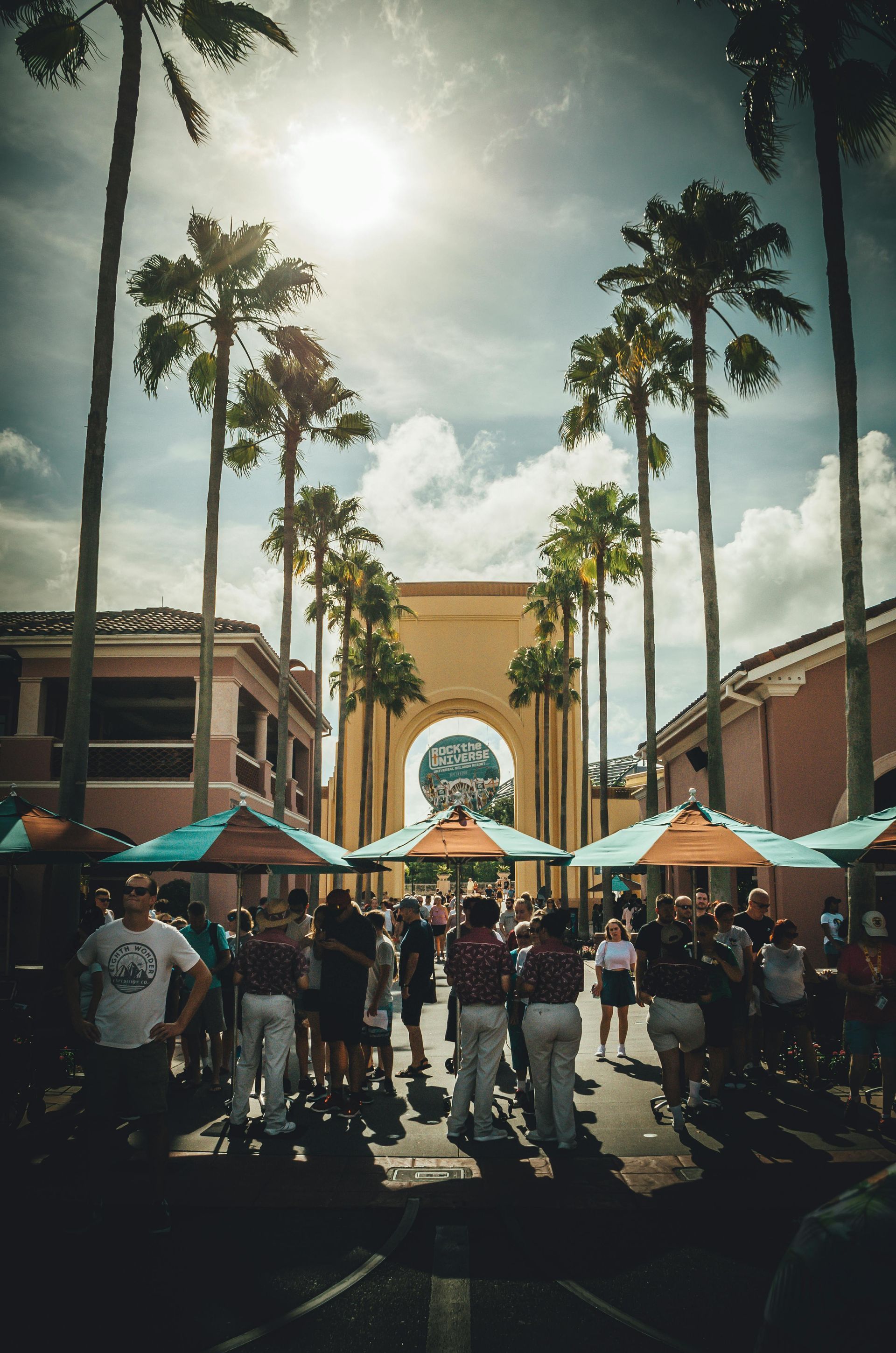 A group of people are standing in front of a building with palm trees and umbrellas.