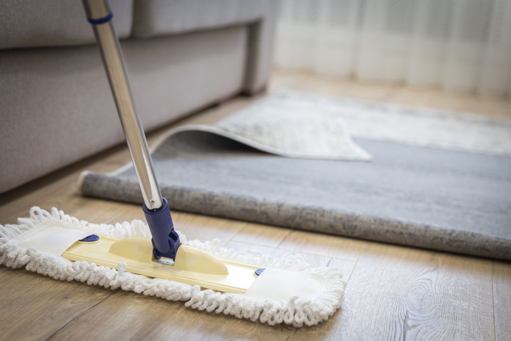 Mop cleaning a light-colored wooden floor, with a gray rug partially rolled up in the background