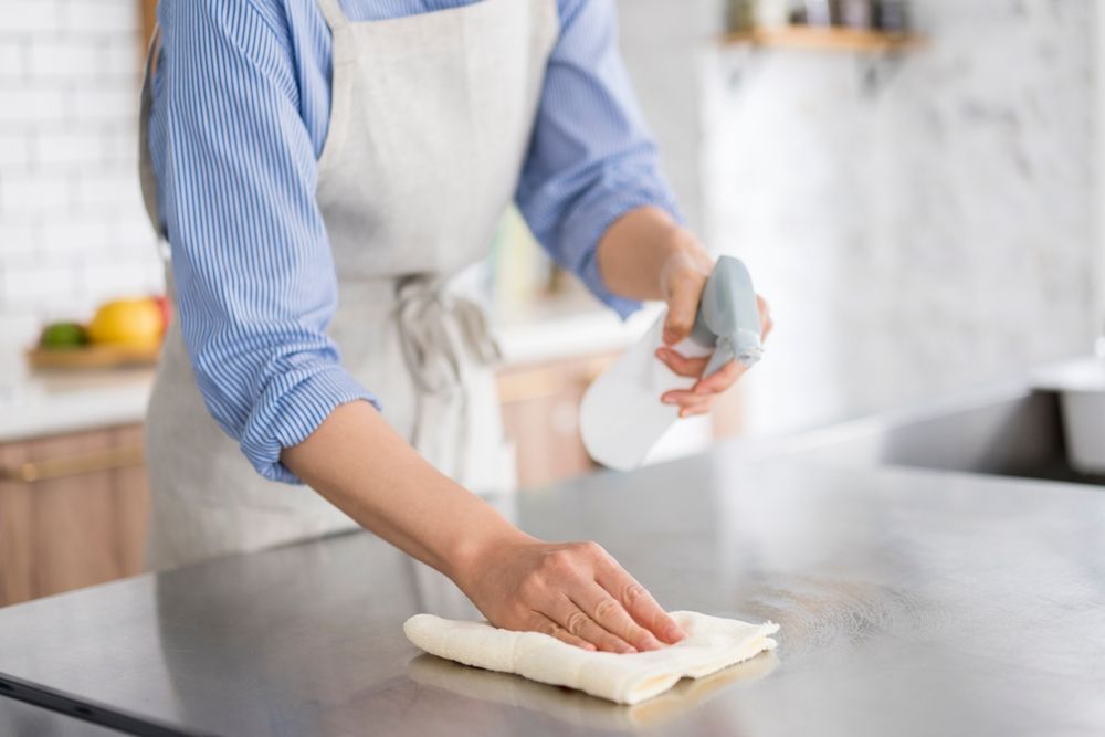 Woman in apron cleaning a stainless steel countertop with spray bottle and cloth in a kitchen