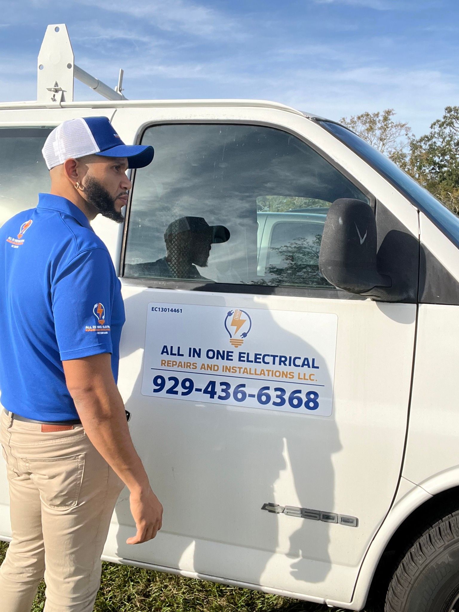 Man in blue shirt and cap stands by a white electrical service van, the company name and phone number are visible.