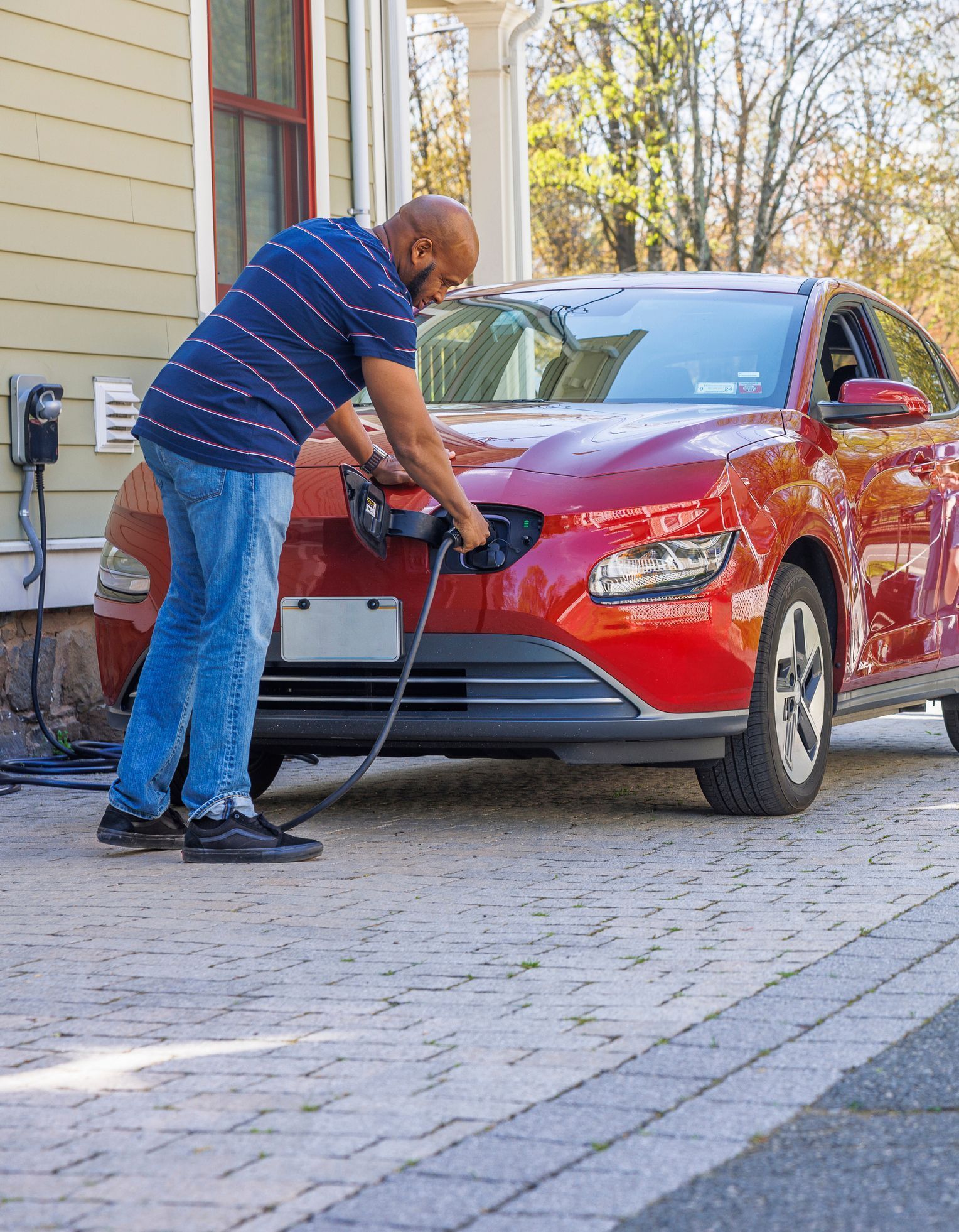 Man charging a red electric car in a driveway with a charger attached to the house.
