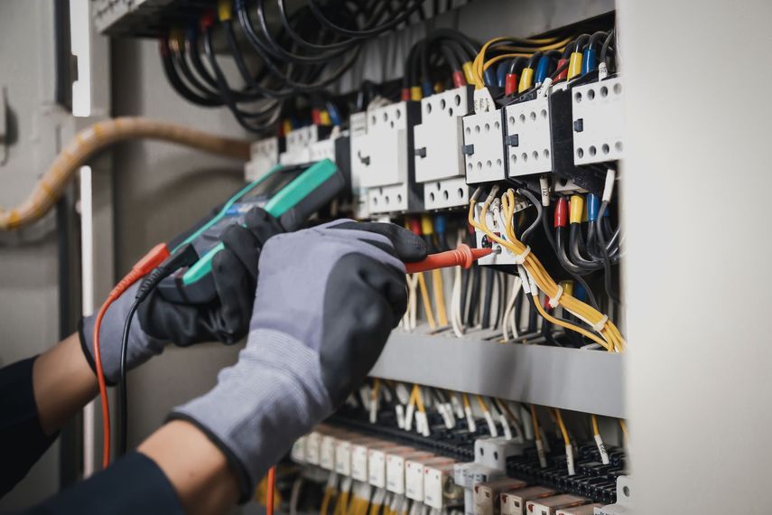 Hands in gloves using a multimeter to test wires in an electrical panel.
