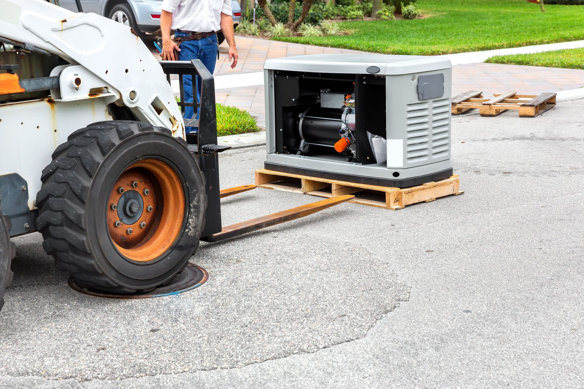 A skid steer forklift moving a generator on a pallet on a residential street, a person in the background.