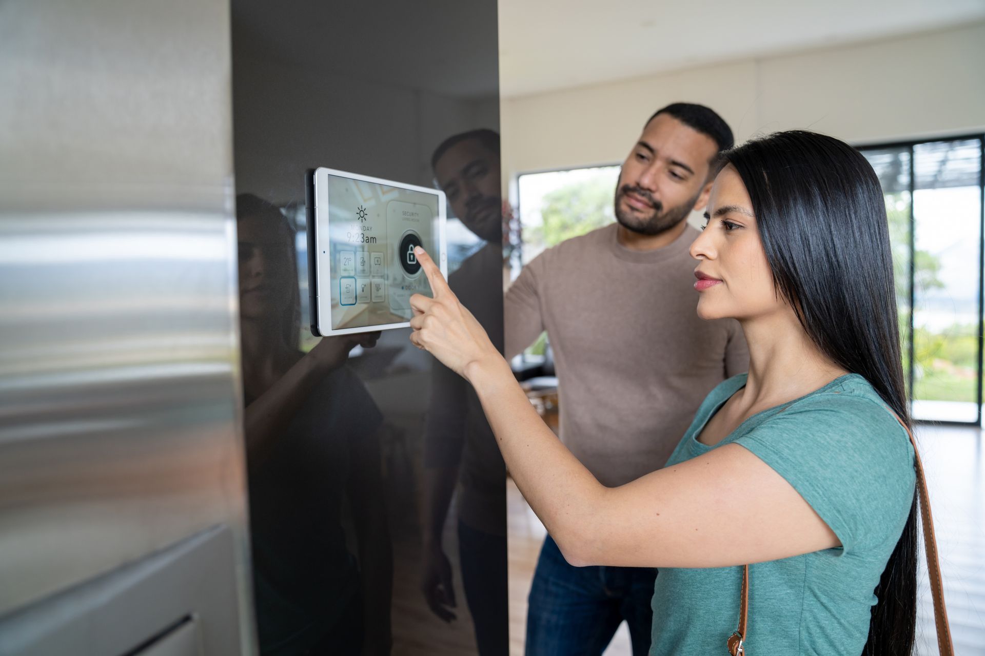 Couple interacting with a smart home control panel on a dark wall. Woman points, man watches. Bright interior, sunny view.