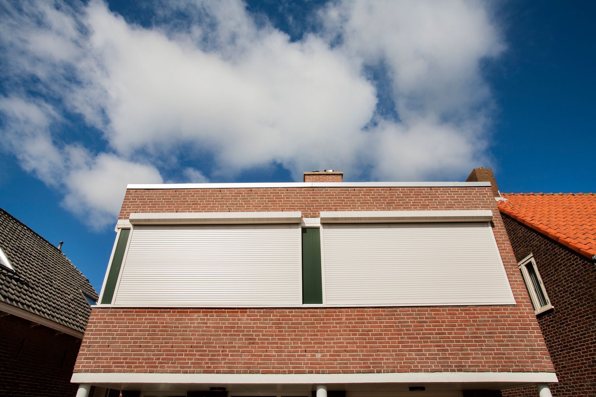 Red brick house with two windows covered by closed white shutters under a blue sky.