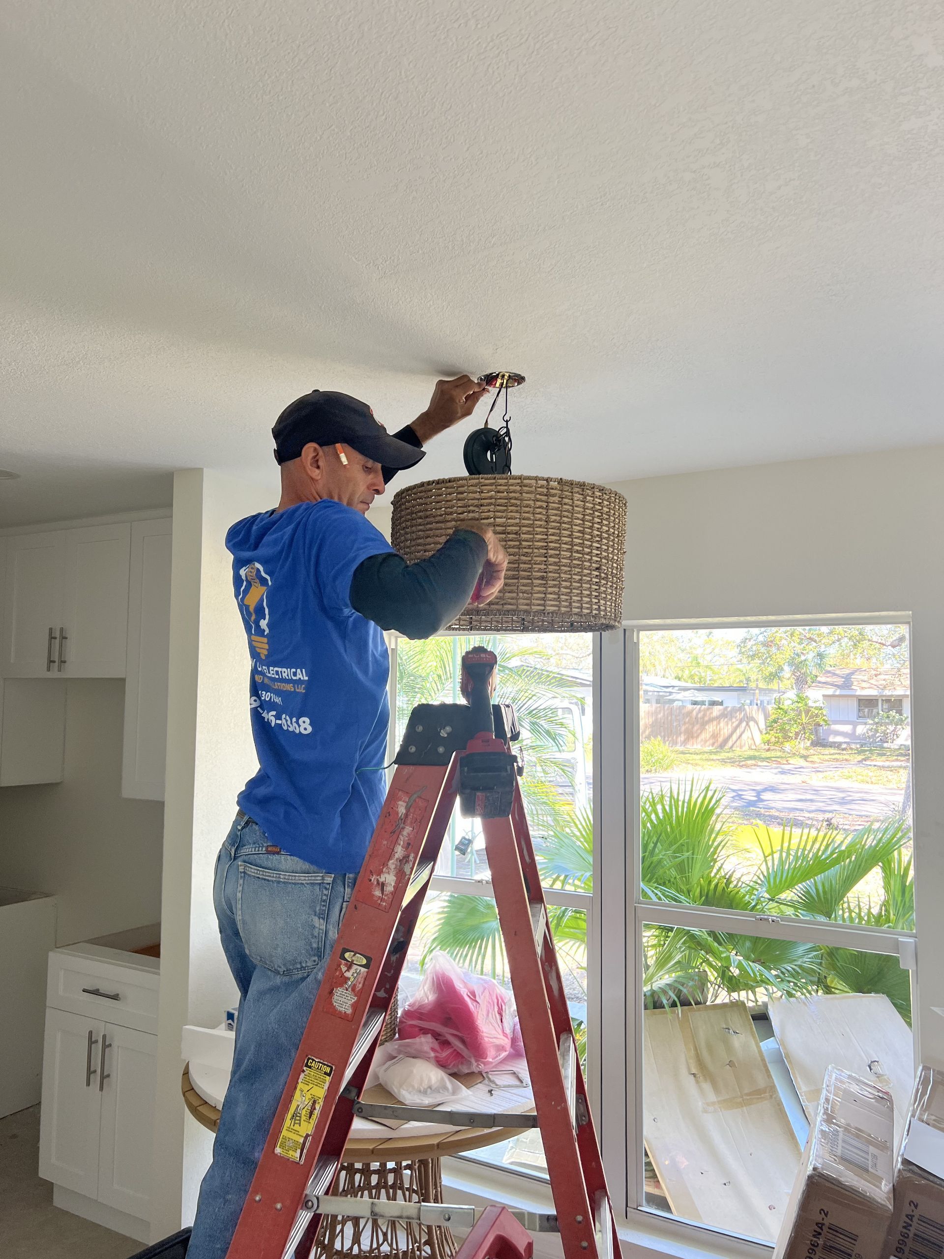 Man on ladder installing a woven light fixture on a white ceiling near a window.