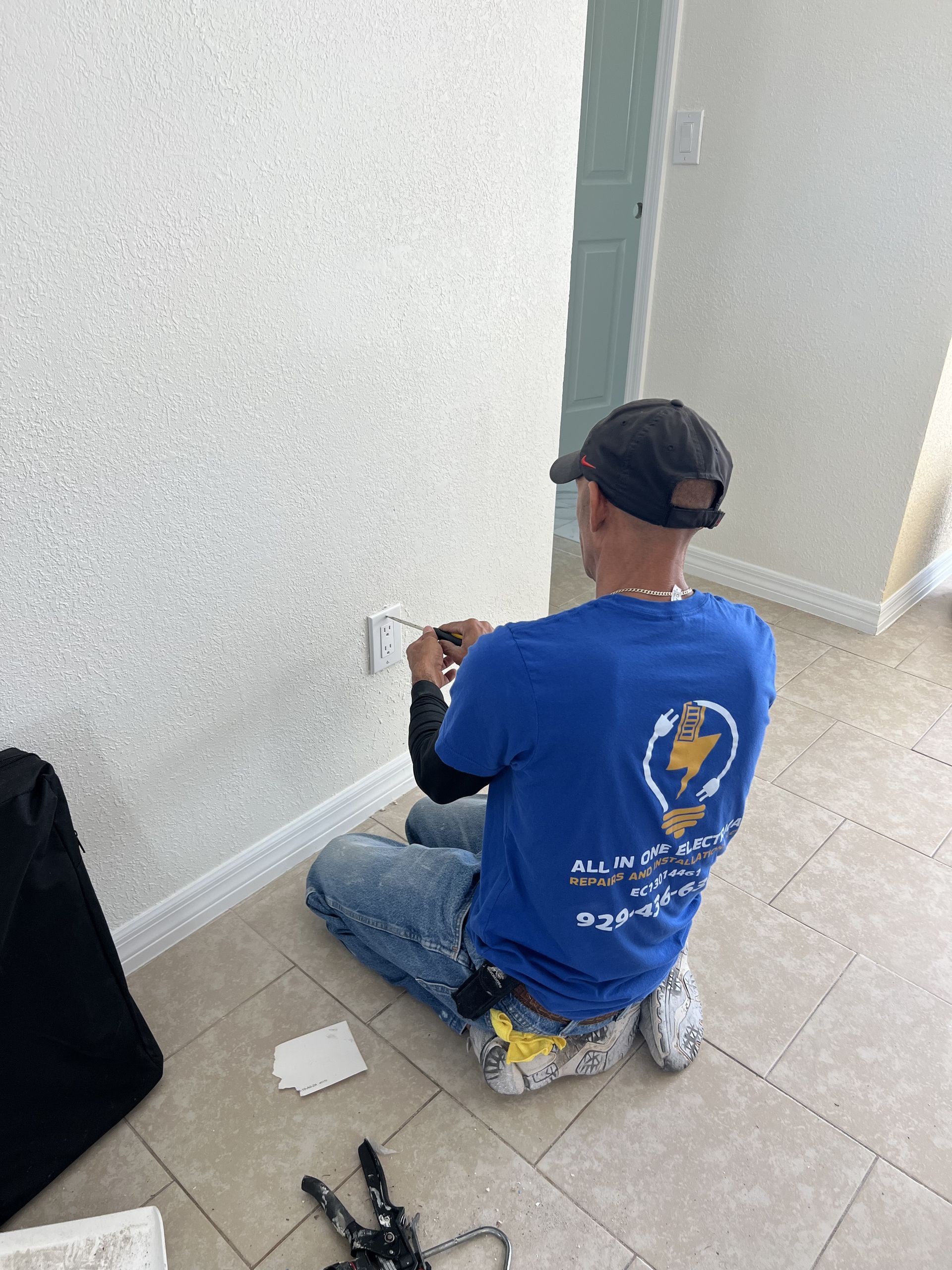 An electrician kneeling, working on a wall outlet in a room with tile flooring and a white wall.