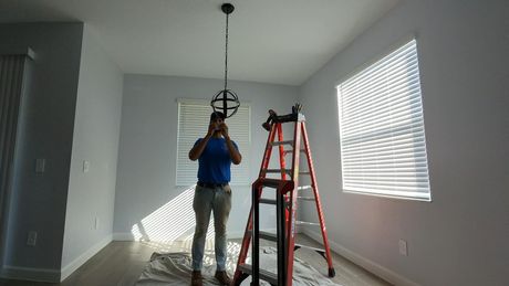 Man installing a light fixture in a room with light gray walls, a ladder, and covered floor.